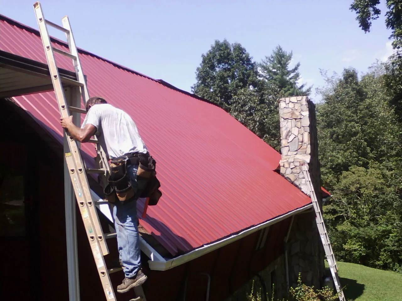 Roofer on a ladder beside a red metal roof and stone chimney — Soffit & Fascia Repair by Roofing Brothers in Ellenville